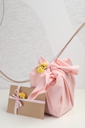 Photo of Furoshiki technique. Gift packed in pink fabric, card, and flowers on white table
