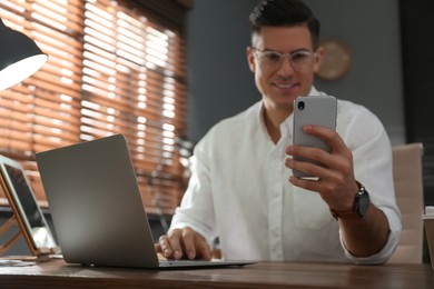 Photo of Freelancer working with laptop and smartphone at table indoors, focus on hand