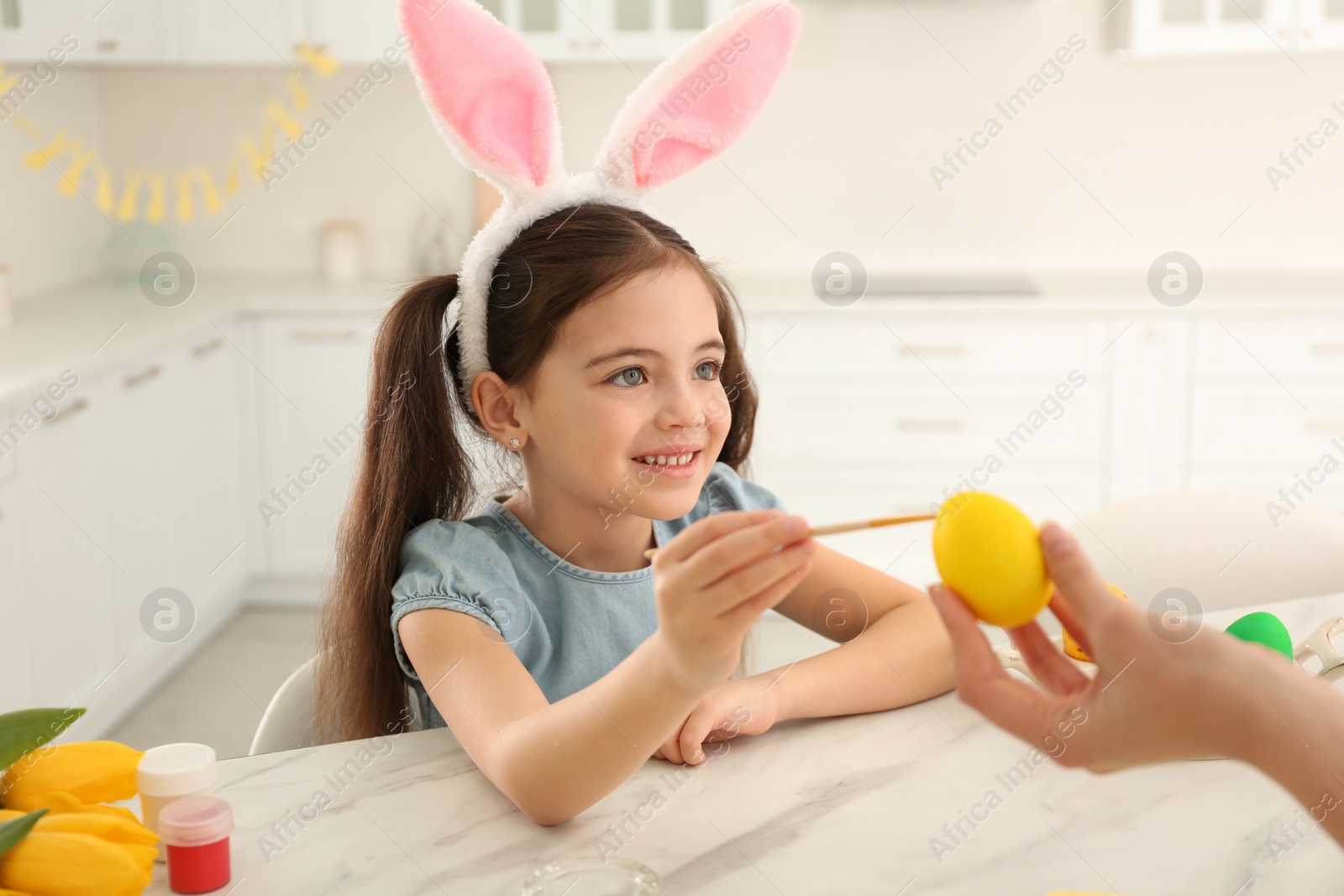 Photo of Happy little girl with bunny ears headband and her mother painting Easter egg in kitchen
