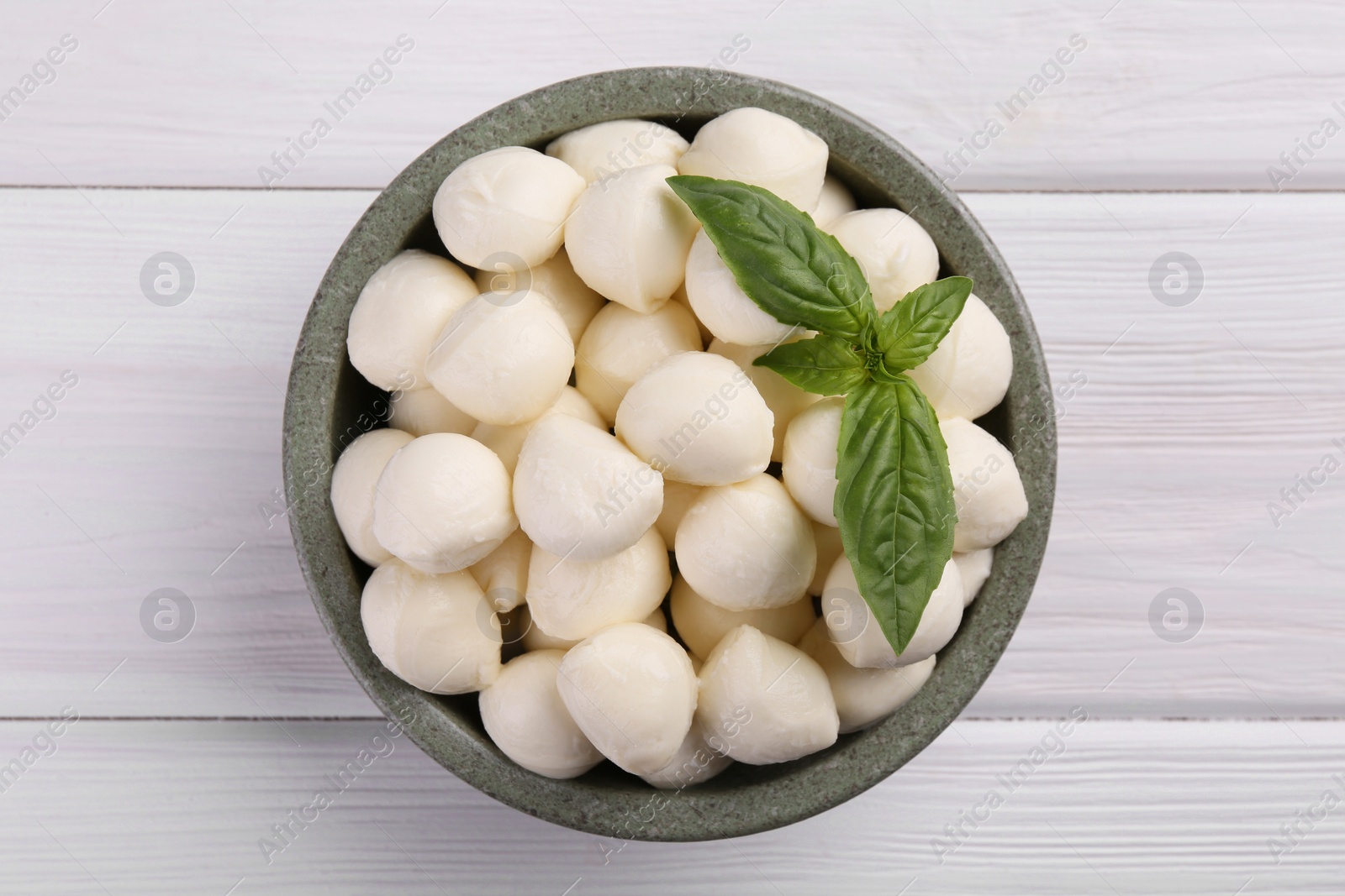 Photo of Tasty mozzarella balls and basil leaves in bowl on white wooden table, top view