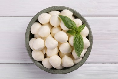 Tasty mozzarella balls and basil leaves in bowl on white wooden table, top view