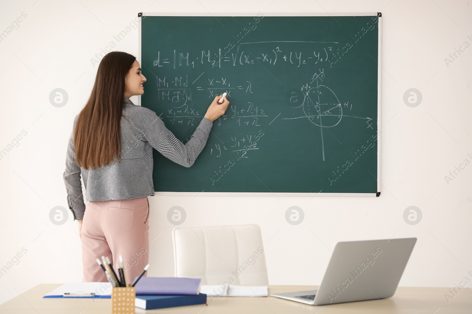 Photo of Young female teacher writing on blackboard in classroom