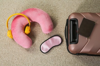 Photo of Pink travel pillow with headphones, suitcase, passport and sleep mask on beige rug, flat lay