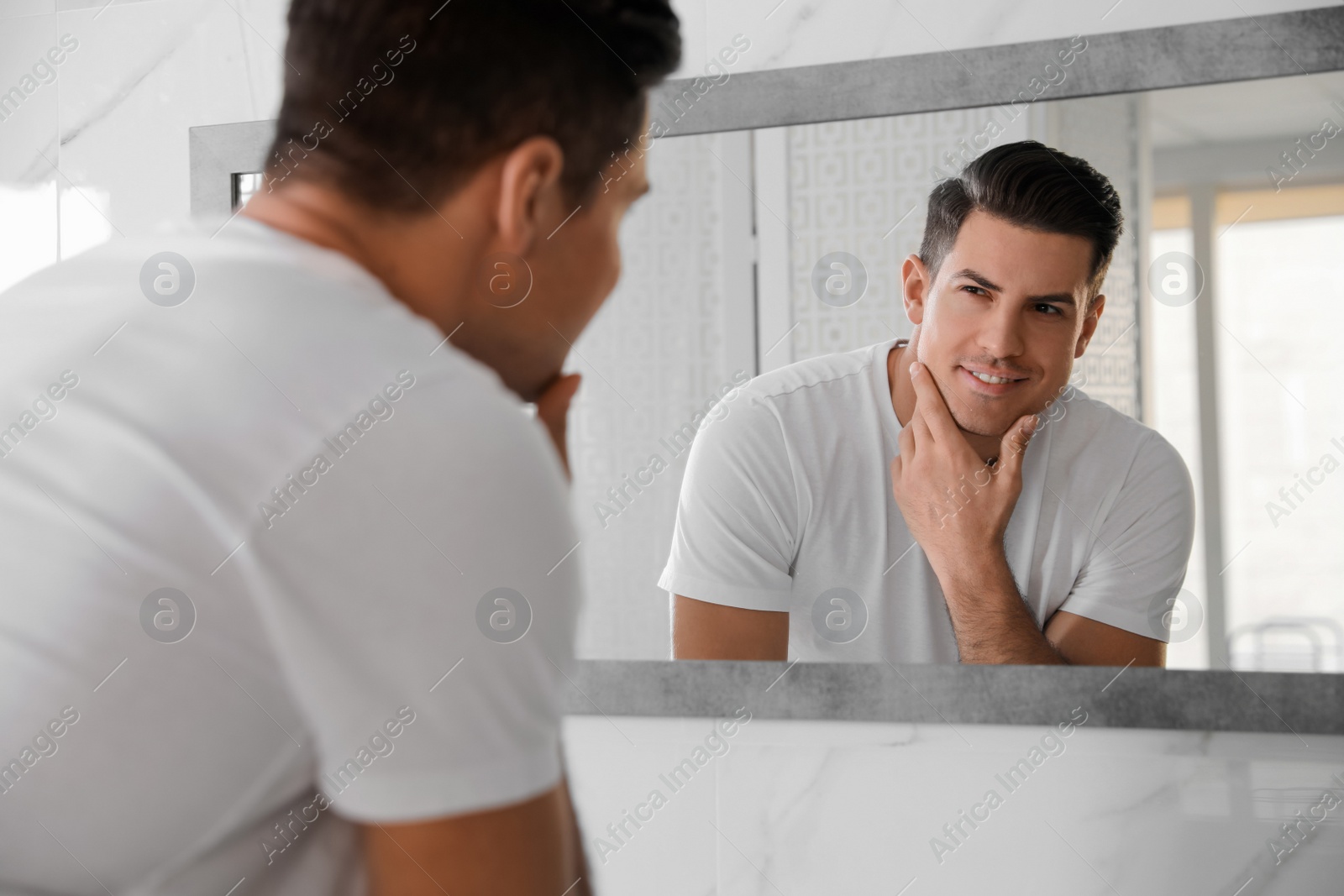 Photo of Handsome man touching his smooth face after shaving near mirror in bathroom