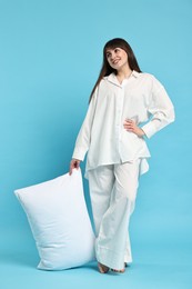 Happy woman in pyjama holding pillow on light blue background