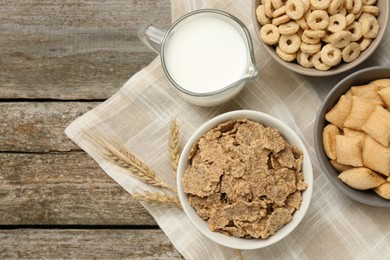 Different breakfast cereals, milk and spikelets on wooden table, flat lay. Space for text