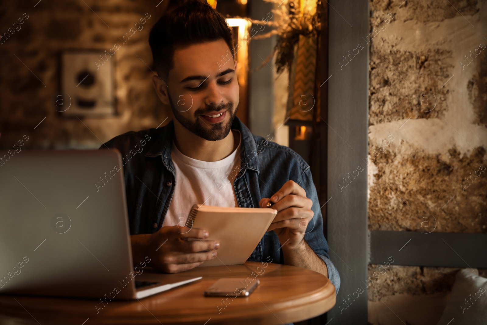 Photo of Young blogger writing in notebook at table indoors