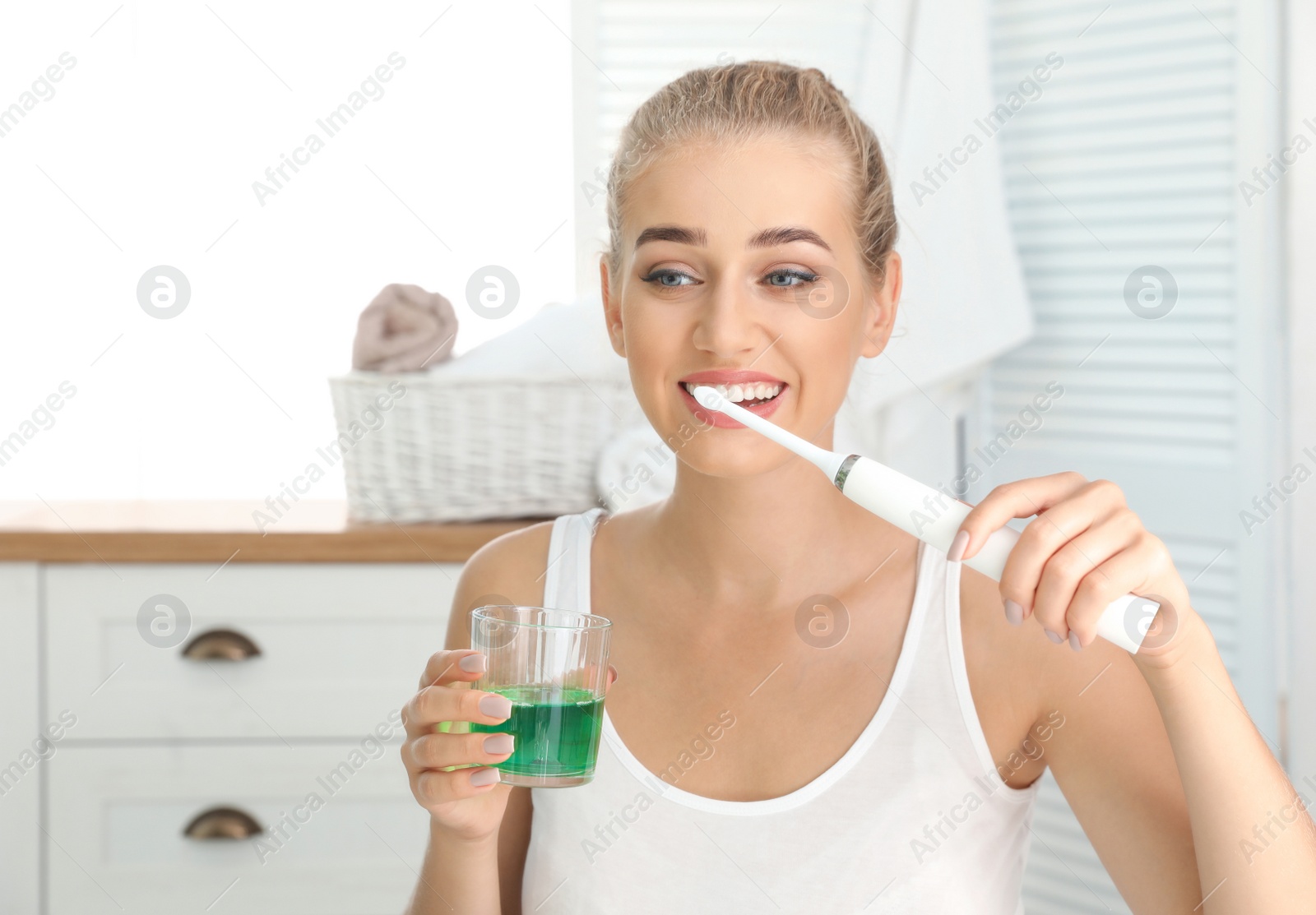 Photo of Woman brushing teeth and holding glass with mouthwash in bathroom