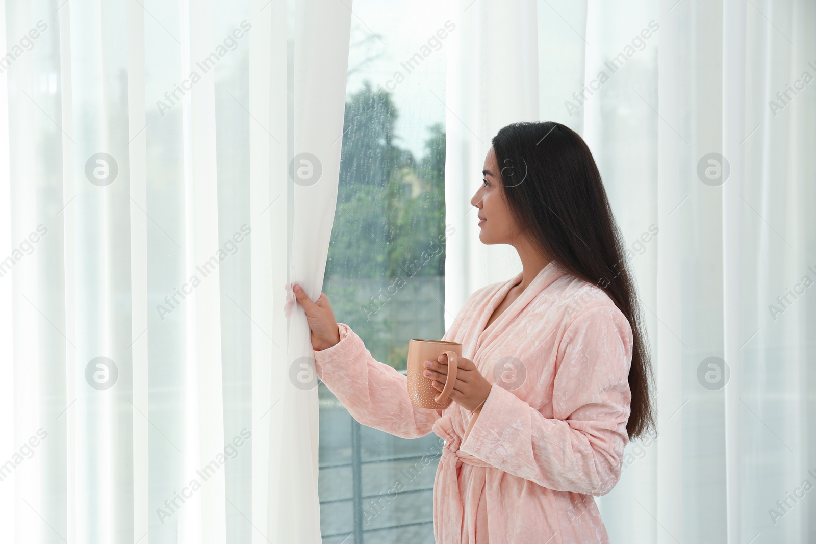Image of Young woman with cup of coffee opening curtains at home. Good morning