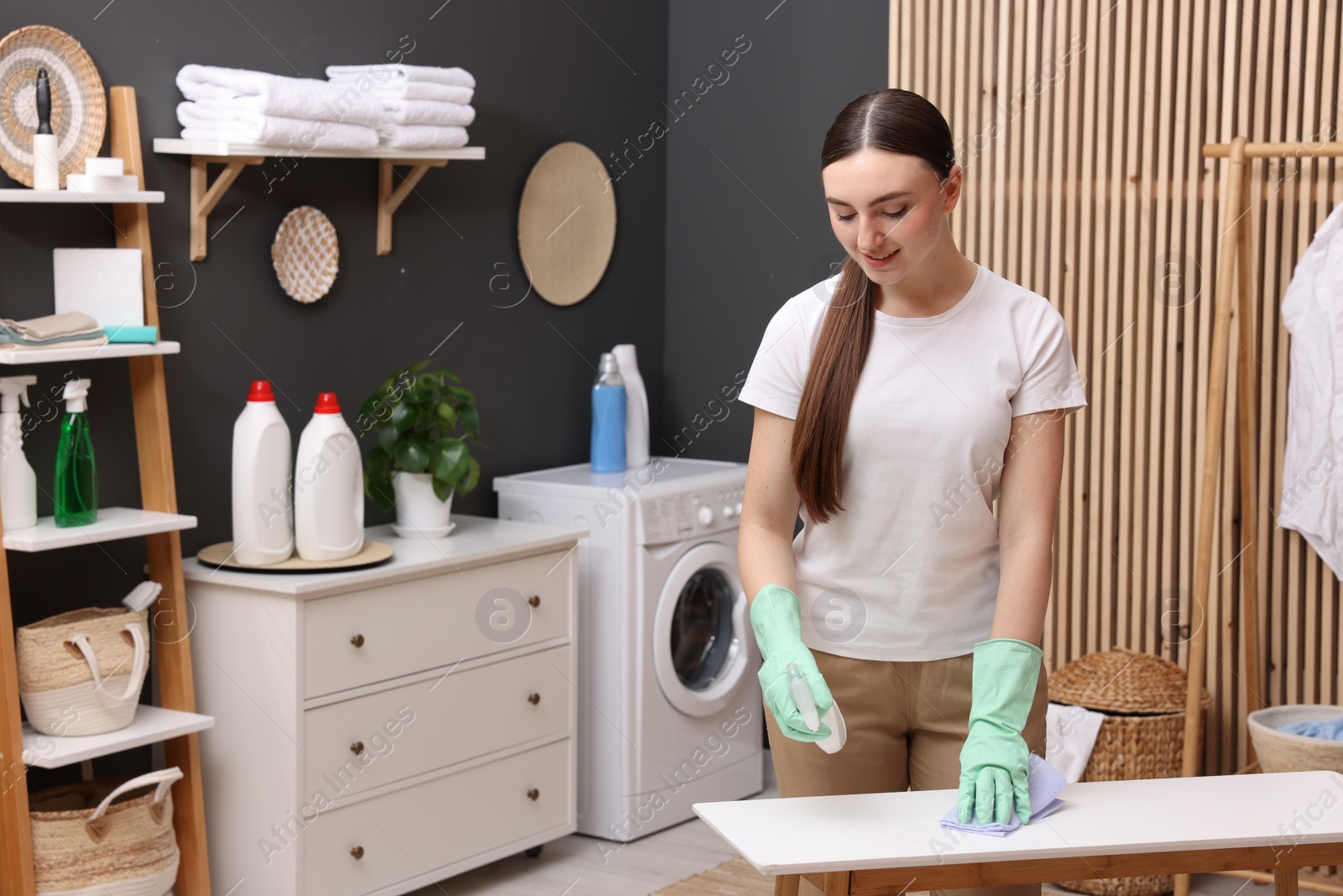 Photo of Beautiful woman with spray bottle and microfiber cloth cleaning white table in laundry room