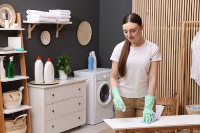 Photo of Beautiful woman with spray bottle and microfiber cloth cleaning white table in laundry room