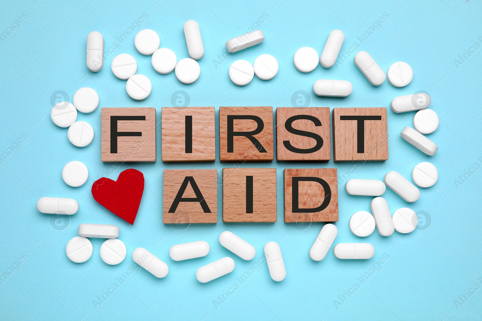 Photo of Words First Aid made of wooden cubes, pills and cardboard heart on light blue background, flat lay