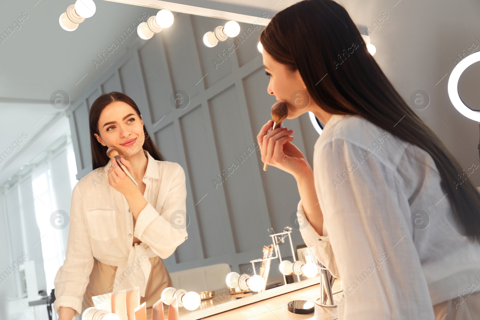 Photo of Young woman applying make up near illuminated mirror indoors