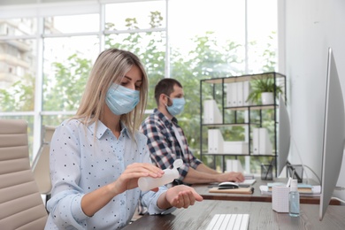 Photo of Office employee in mask applying hand sanitizer at workplace