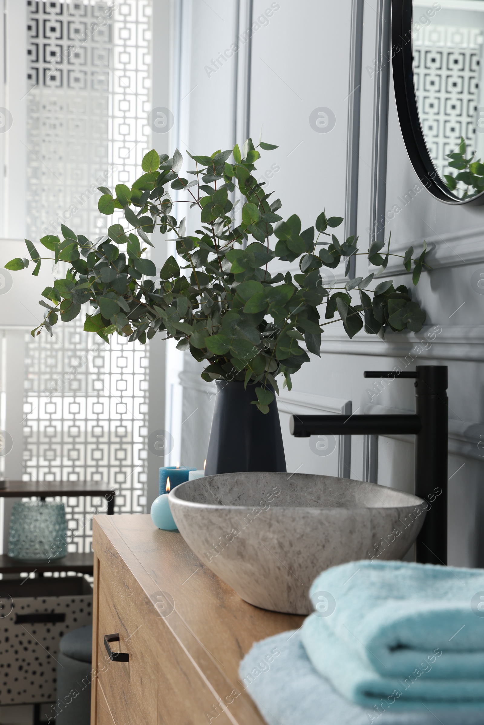 Photo of Eucalyptus branches near vessel sink, burning candles and towels on bathroom vanity. Interior design