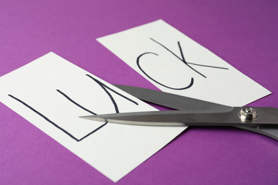 Photo of Cut paper with word LUCK and scissors on purple background, closeup