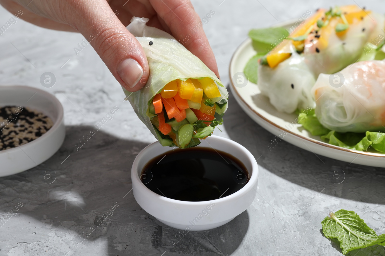 Photo of Woman dipping delicious spring roll into soy sauce at grey textured table, closeup