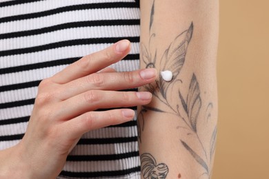 Tattooed woman applying cream onto her arm on beige background, closeup