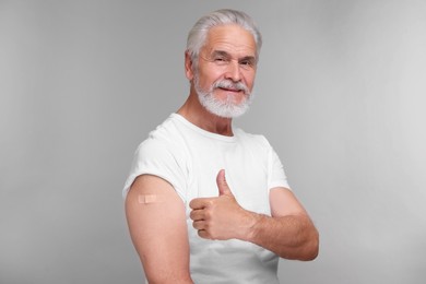 Photo of Senior man with adhesive bandage on his arm after vaccination showing thumb up against light grey background