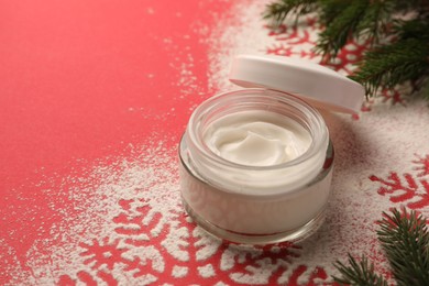 Winter skin care. Hand cream near snowflake silhouettes made with artificial snow and fir branches on red background, closeup