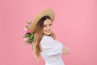 Happy young woman in straw hat holding bouquet of beautiful tulips on pink background