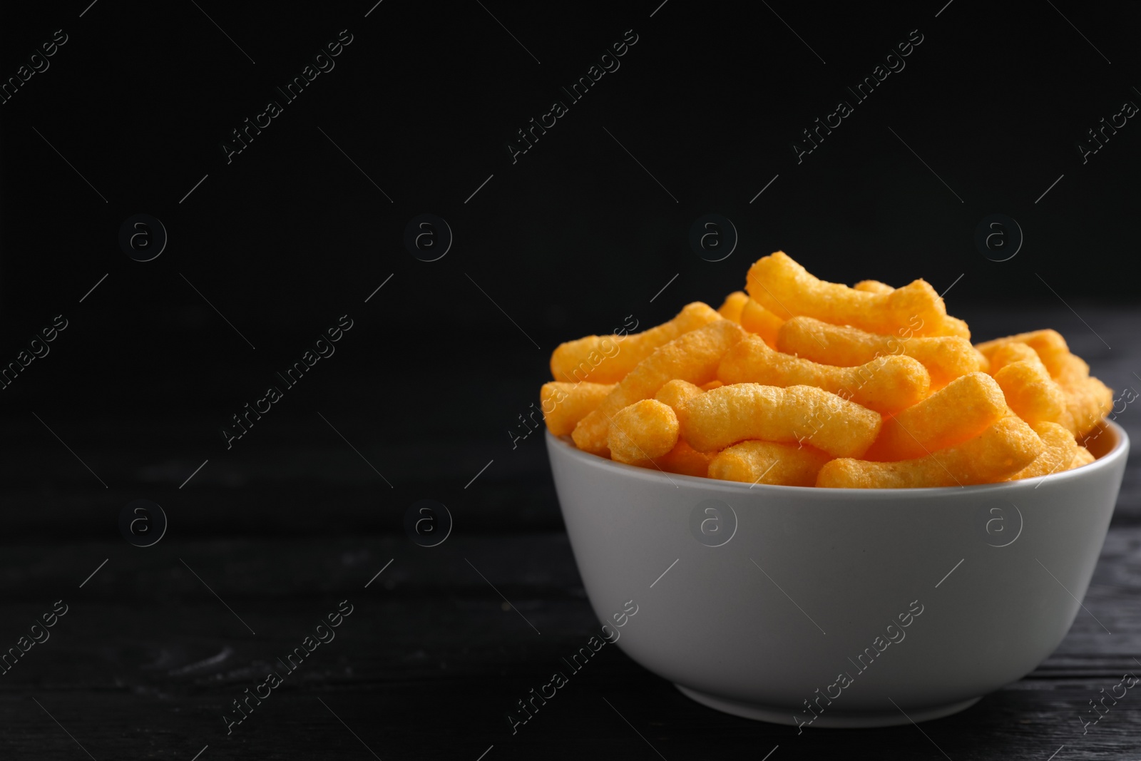 Photo of Bowl with crunchy cheesy corn snack on black wooden table. Space for text