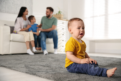 Photo of Unhappy baby sitting alone on floor while parents spending time with his elder brother at home. Jealousy in family