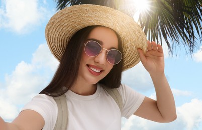 Image of Smiling young woman in sunglasses and straw hat taking selfie under palm tree on sunny day