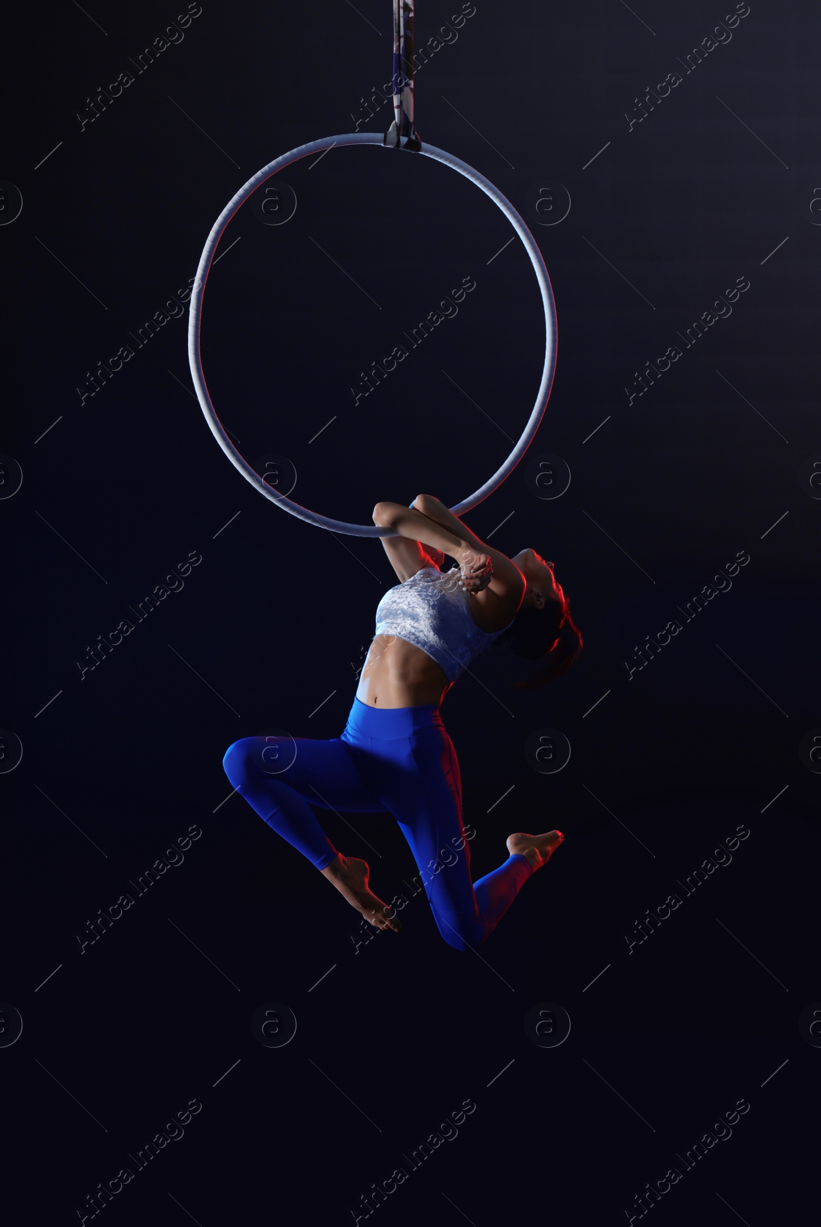 Photo of Young woman performing acrobatic element on aerial ring against dark background