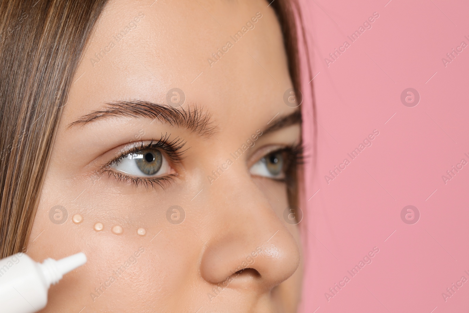 Photo of Young woman applying cream under eyes on pink background, closeup. Space for text