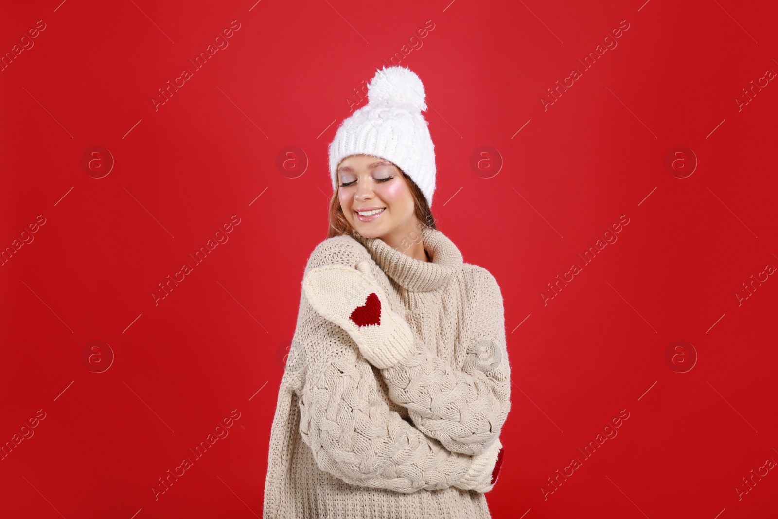 Photo of Young woman in warm sweater, mittens and hat on red background. Winter season