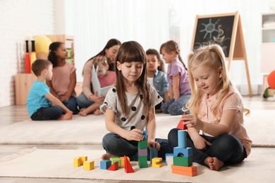 Photo of Cute girls playing with building blocks on floor while kindergarten teacher reading book to other children indoors