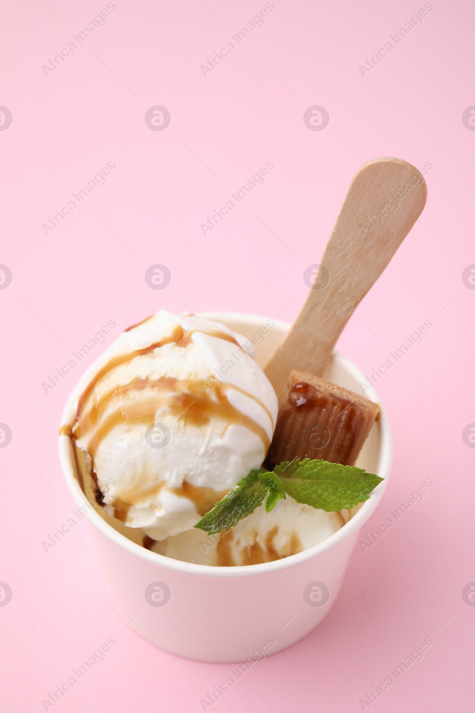 Photo of Tasty ice cream with caramel sauce, mint leaves and candy in paper cup on pink table