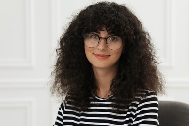 Portrait of beautiful woman with curly hair sitting on armchair indoors. Attractive lady looking at camera