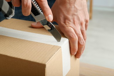 Photo of Man using utility knife to open parcel indoors, closeup