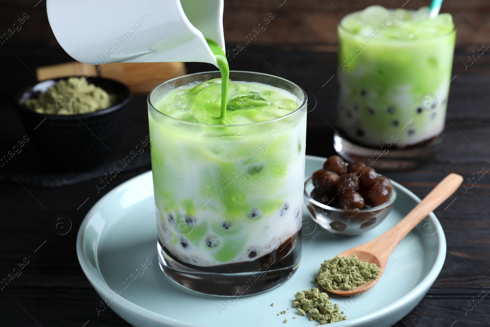 Photo of Pouring green matcha into glass with milk bubble tea at black wooden table, closeup