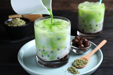 Photo of Pouring green matcha into glass with milk bubble tea at black wooden table, closeup