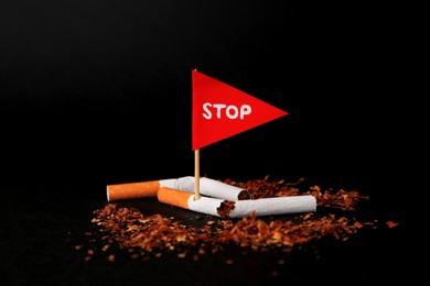 Photo of Broken cigarettes and red flag with word Stop on black background, closeup. Quitting smoking concept