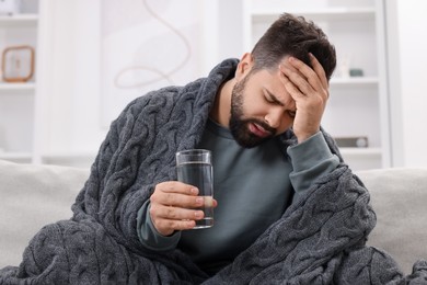 Photo of Man with glass of water suffering from headache on sofa at home