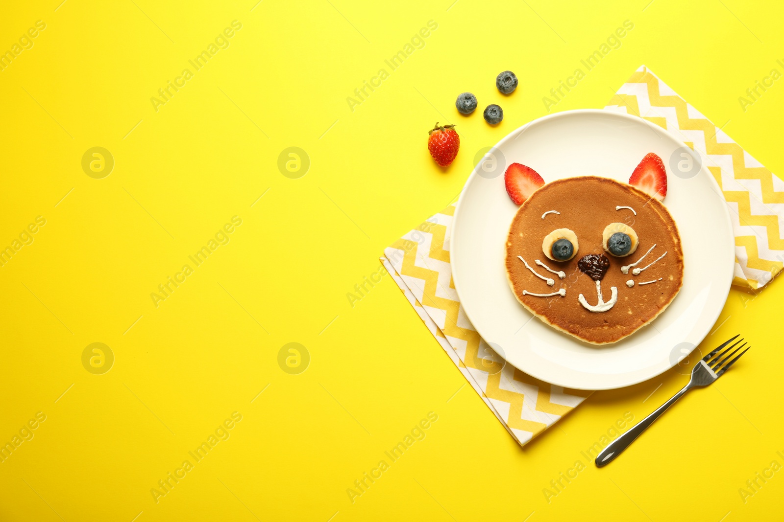 Photo of Creative serving for kids. Plate with cute cat made of pancakes, berries, cream, banana and chocolate paste on yellow background, flat lay. Space for text