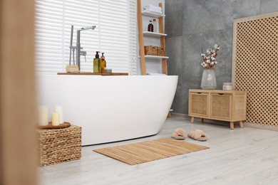 Photo of Stylish bathroom interior with bamboo bath mat and white tub