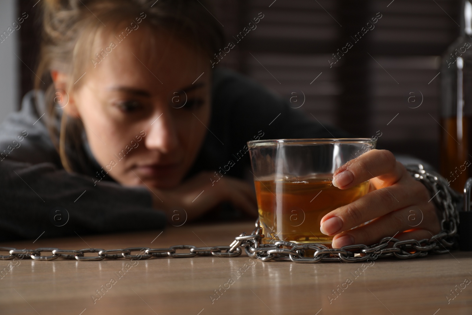 Photo of Alcohol addiction. Woman chained with glass of liquor at wooden table in room, focus on hand