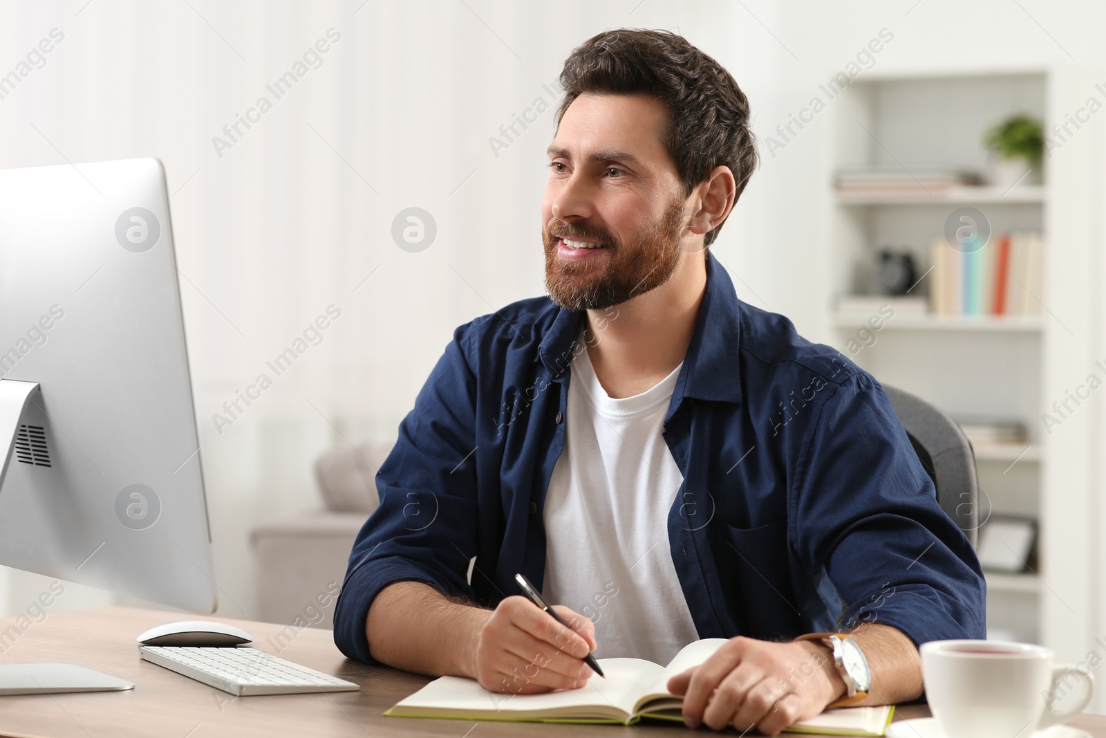 Photo of Home workplace. Happy man taking notes while working with computer at wooden desk in room
