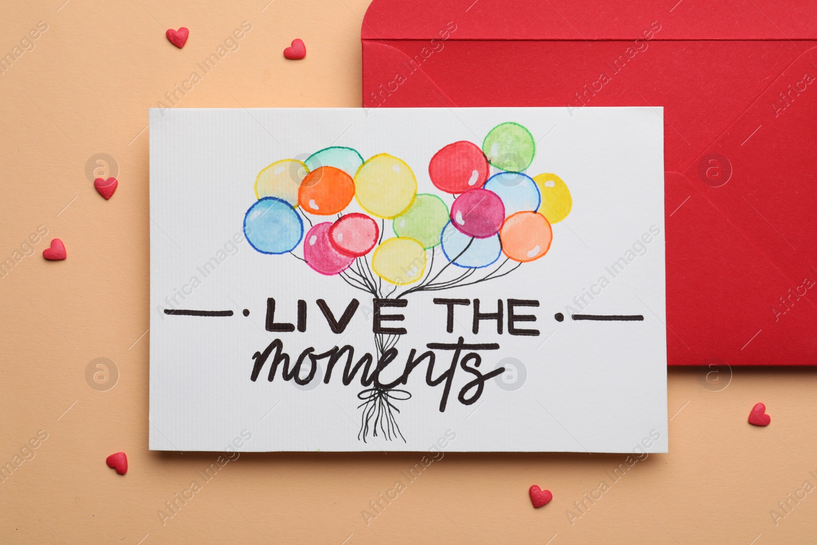 Photo of Card with phrase Live The Moments and red envelope on beige background, flat lay