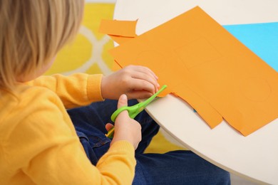 Photo of Little boy cutting orange paper at desk in room, closeup. Home workplace