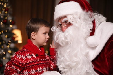 Photo of Little boy whispering in Santa Claus' ear near Christmas tree indoors