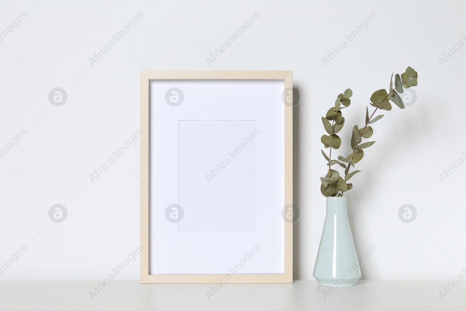 Photo of Empty photo frame and vase with decorative eucalyptus leaves on white table. Mockup for design