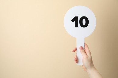 Photo of Woman holding auction paddle with number 10 on beige background, closeup. Space for text