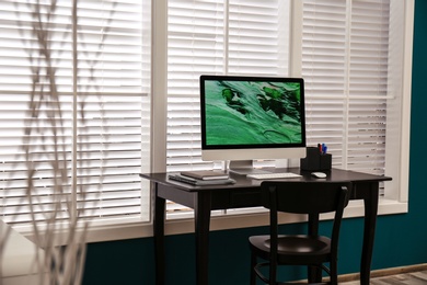 Comfortable workplace near window with white horizontal blinds in room