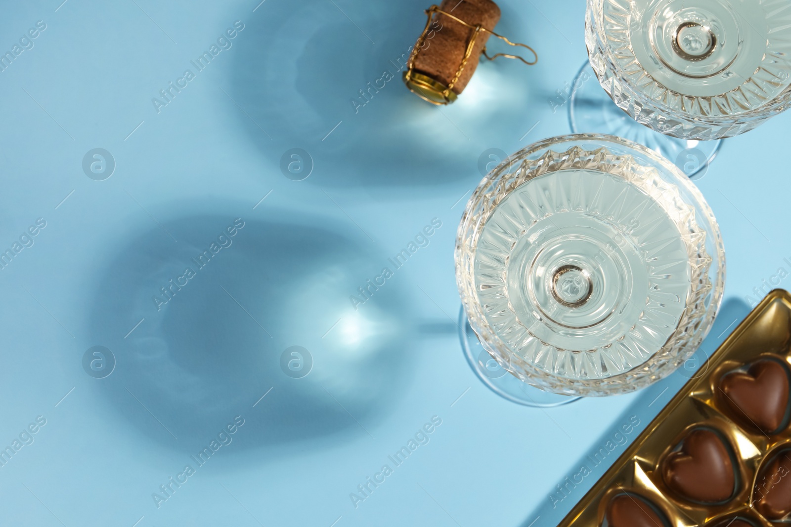 Photo of Glasses of expensive white wine, cork and heart shaped chocolate candies on light blue background, flat lay. Space for text